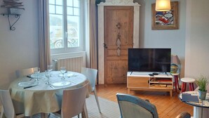 Comedor con una mesa redonda dispuesta para cuatro personas, un televisor y una pequeña mesa con libros y una planta.
