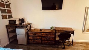 Una habitación compacta con un escritorio de madera, un refrigerador pequeño, un microondas, una silla de oficina negra y un televisor montado en la pared.