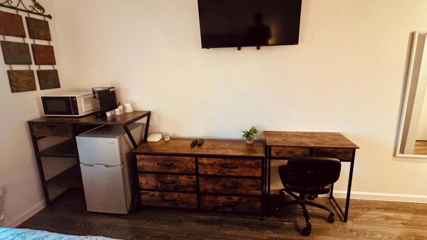 A compact room with a wooden desk, a small refrigerator, a microwave, a black office chair, and a TV mounted on the wall. - 24 - Lovely stays start here (Challenge-Brownsville)