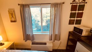 A room with a bed, a bedside lamp, a window with curtains, an air conditioner, a microwave, and a refrigerator. - 24 - Lovely stays start here (Challenge-Brownsville)