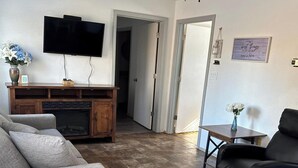 A living room with a flat-screen TV, a wooden entertainment unit, a fireplace, a sofa, a coffee table, and a door leading to another room. - 11 - 1 king, 1 full, 1 queen, 2br, 1ba suite (Challenge-Brownsville)