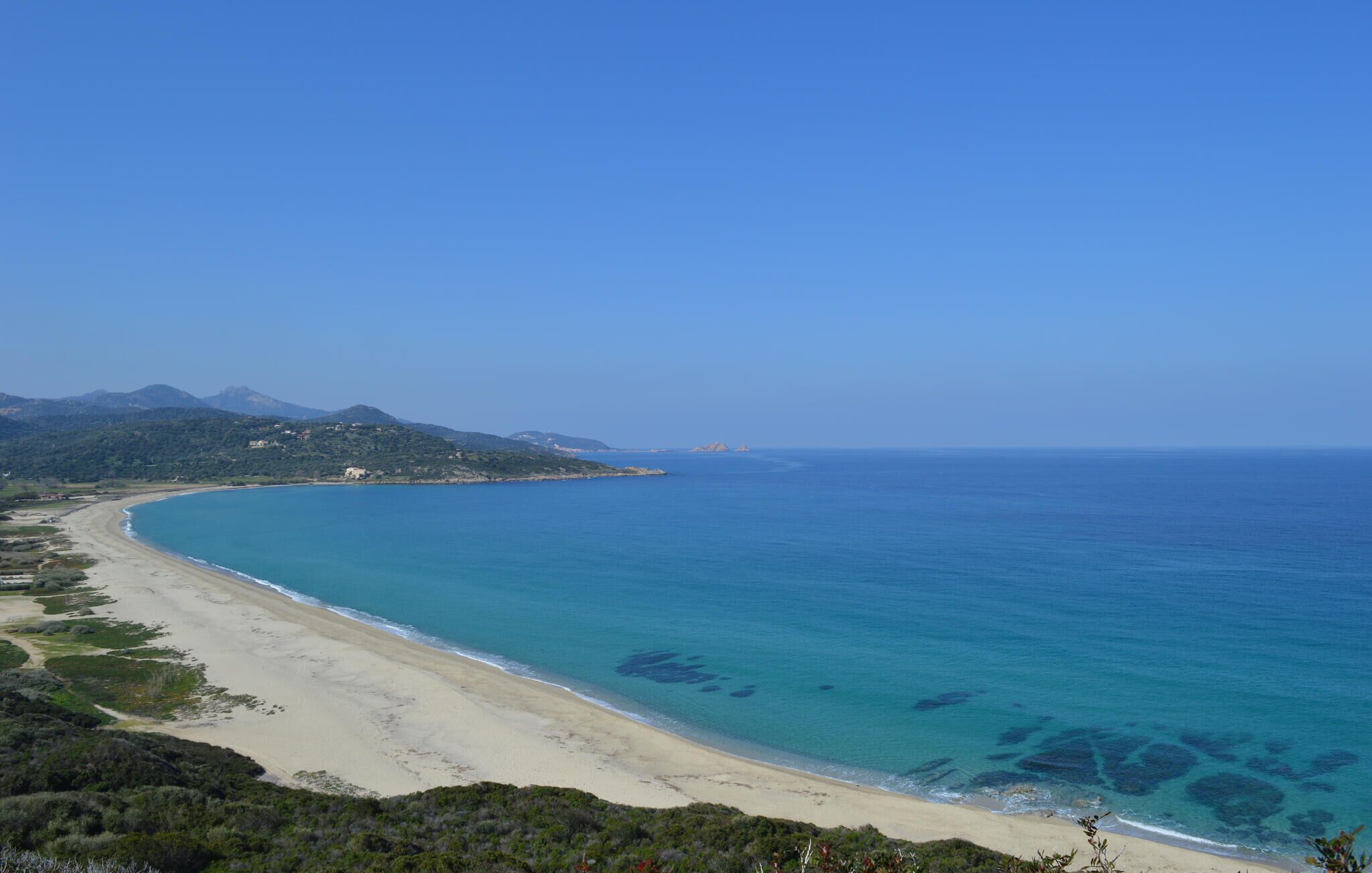 A coastal landscape with a sandy beach, clear blue sea, and distant mountains.