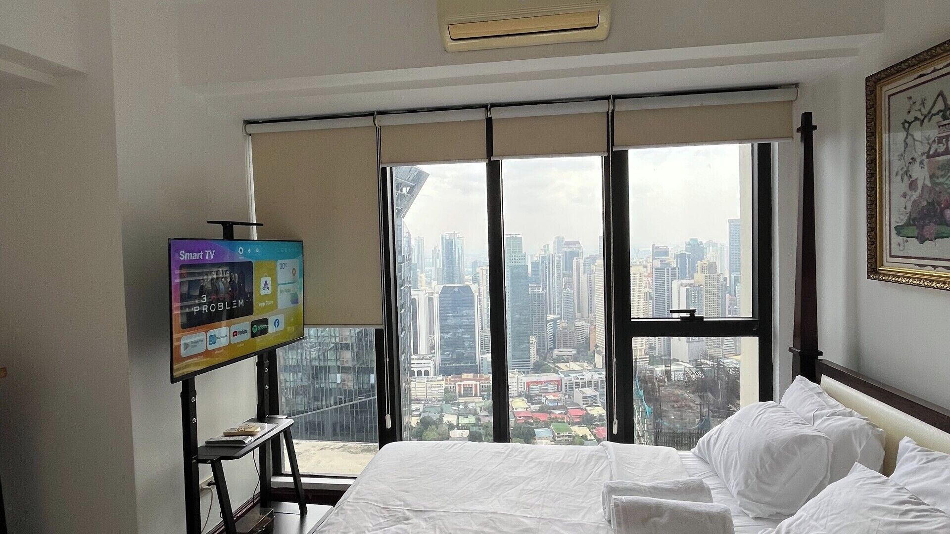Een hotelkamer met een bed, een televisie, een raam met uitzicht over de stad en een ingelijst kunstwerk aan de muur.