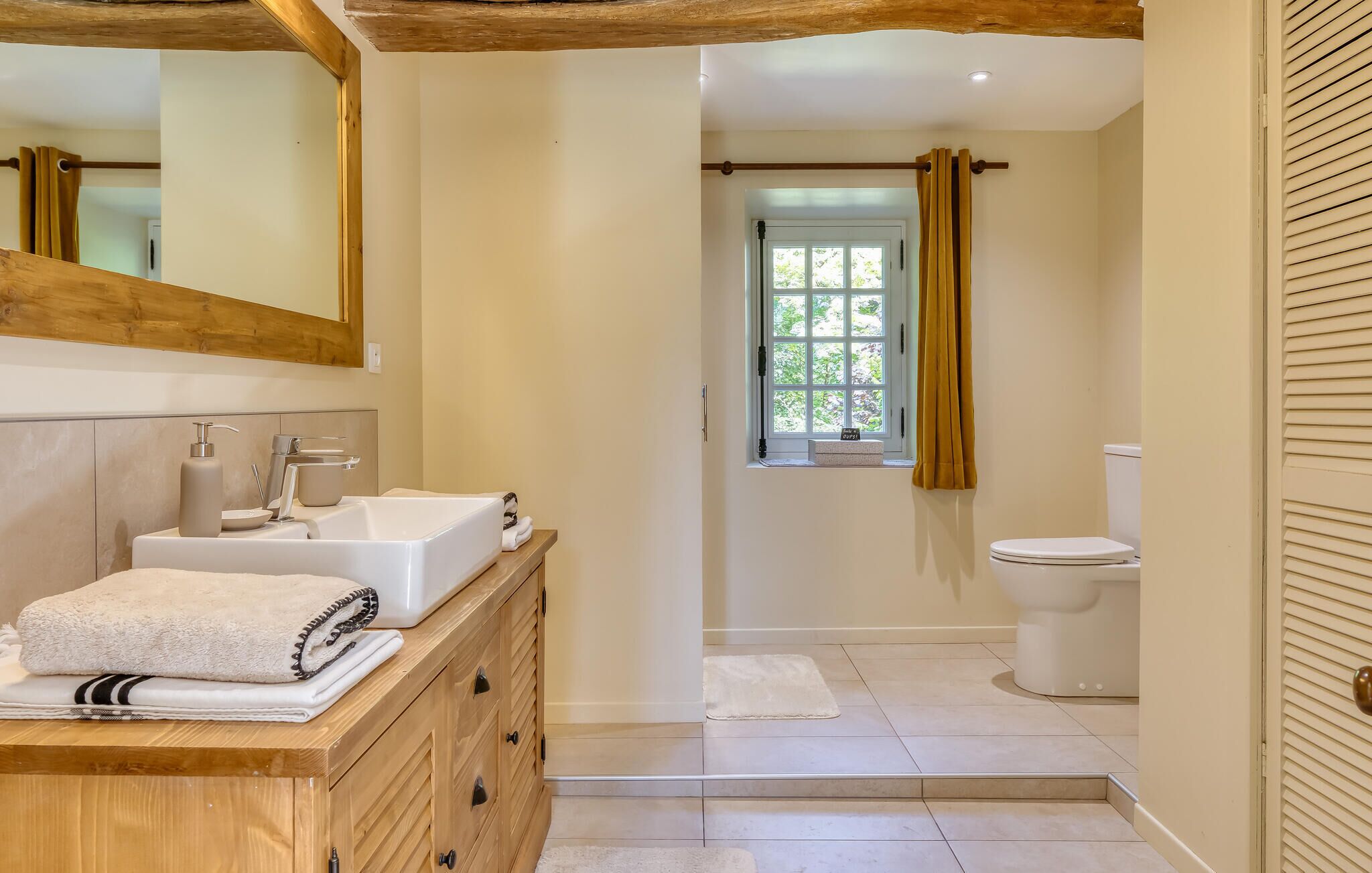 Un baño con tocador de madera, un lavabo, un inodoro y una ventana con cortinas.