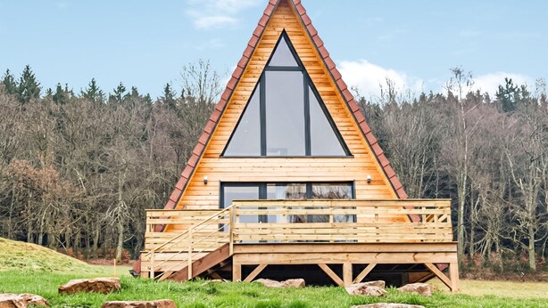 Une cabane en bois avec un toit triangulaire et de grandes fenêtres, entourée d’une forêt.