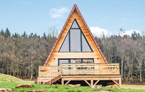 Une cabane en bois avec un toit triangulaire et de grandes fenêtres, entourée d’une forêt.