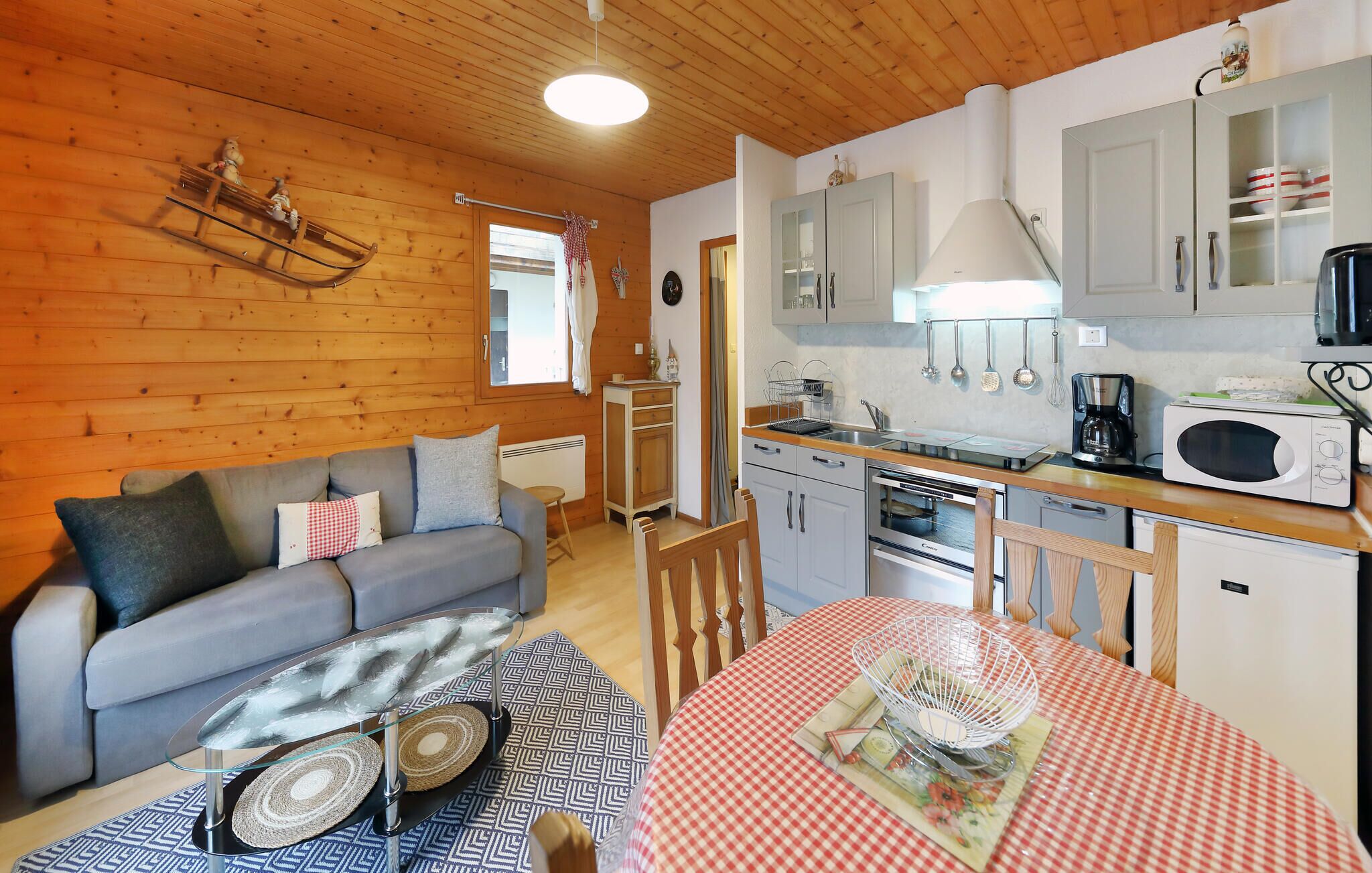 Une pièce confortable, aménagée comme un chalet, avec un canapé, une table à manger recouverte d’une nappe à carreaux, une cuisine équipée d’un four à micro-ondes et un luminaire suspendu.