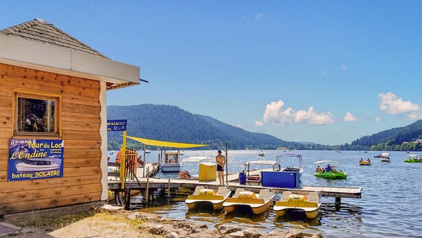 A wooden building with a sign for 'L'Ondine' and 'bateau SOLAIRE'. There are several small boats docked in the water, and a few people are visible on the dock. - Amazing apartment in Gerardmer with lake view (Gerardmer)