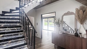 Una escalera con peldaños de mármol negro y barandilla geométrica, que conduce a una habitación con suelo de madera, un espejo grande y un jarrón con plantas secas.