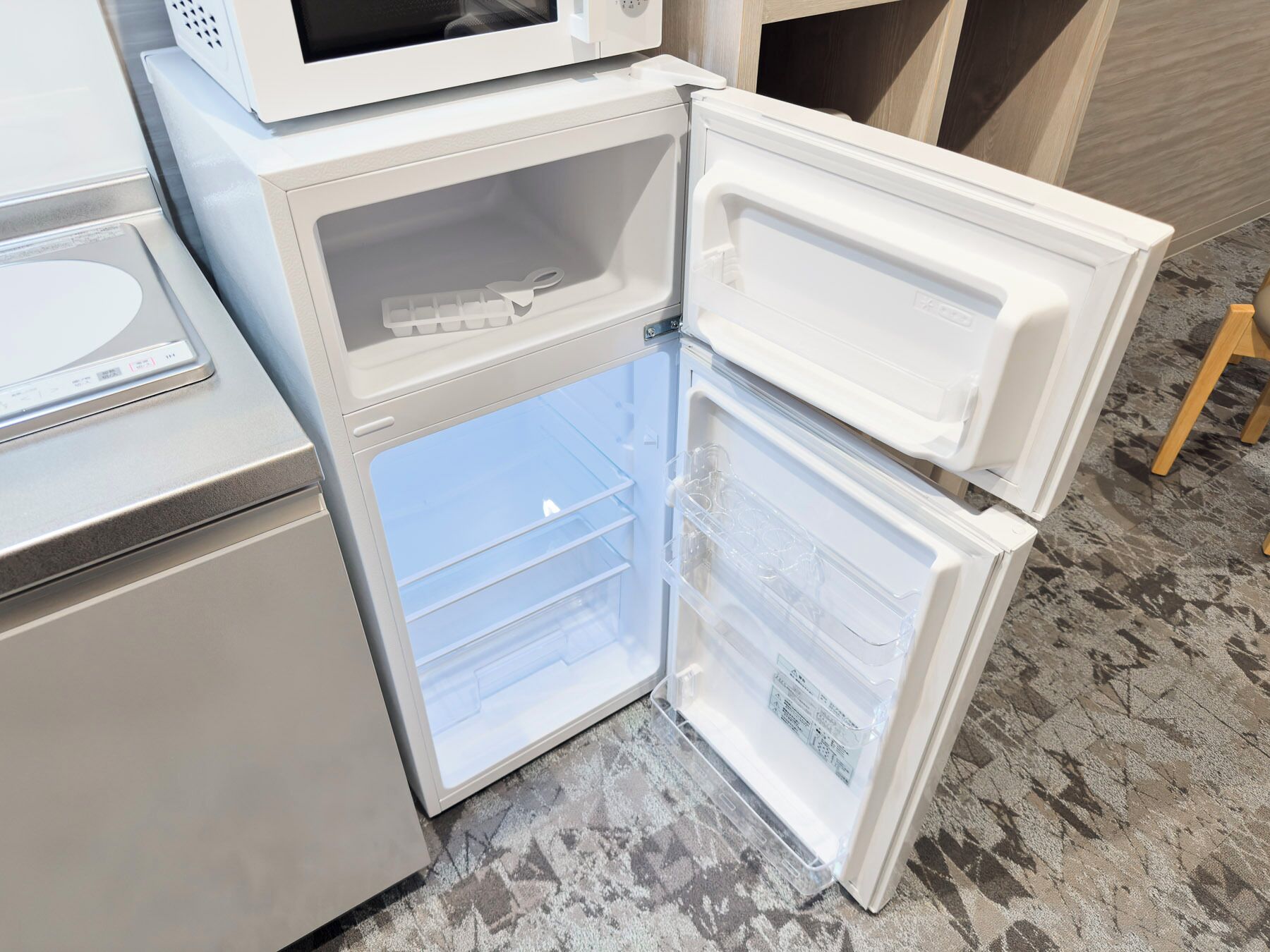Un réfrigérateur blanc doté d’un compartiment congélateur, un four à micro-ondes placé sur le dessus, et un petit tabouret en bois à proximité.