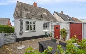 A house with a grey exterior, a tiled roof, a red door, and a small garden area with a table and chairs.