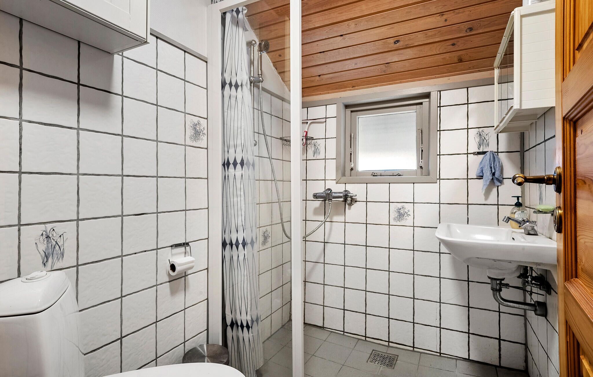 A bathroom with white tiled walls, a wooden ceiling, a shower with a curtain, a window, and a sink with a soap dispenser.
