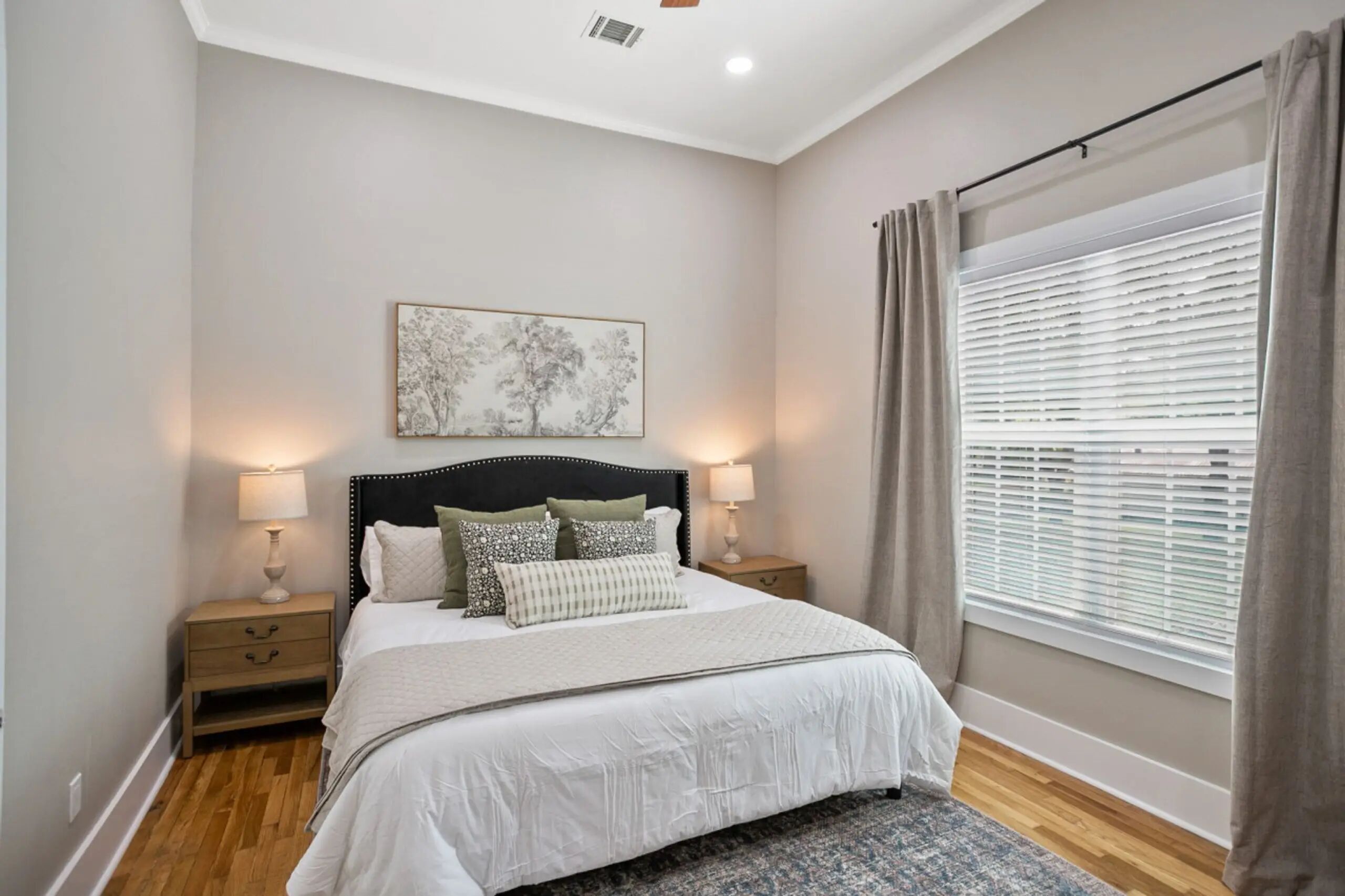 A bedroom with a bed, bedside tables, a nightstand, a window with blinds, and a ceiling light.