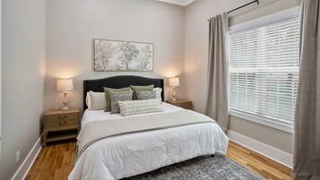 A bedroom with a bed, bedside tables, a nightstand, a window with blinds, and a ceiling light.