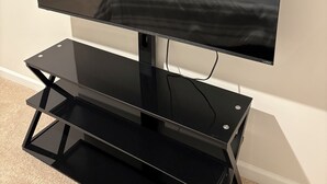 Un téléviseur à écran plat, posé sur un meuble noir, dans une pièce où l’on aperçoit un lit et une porte au fond.