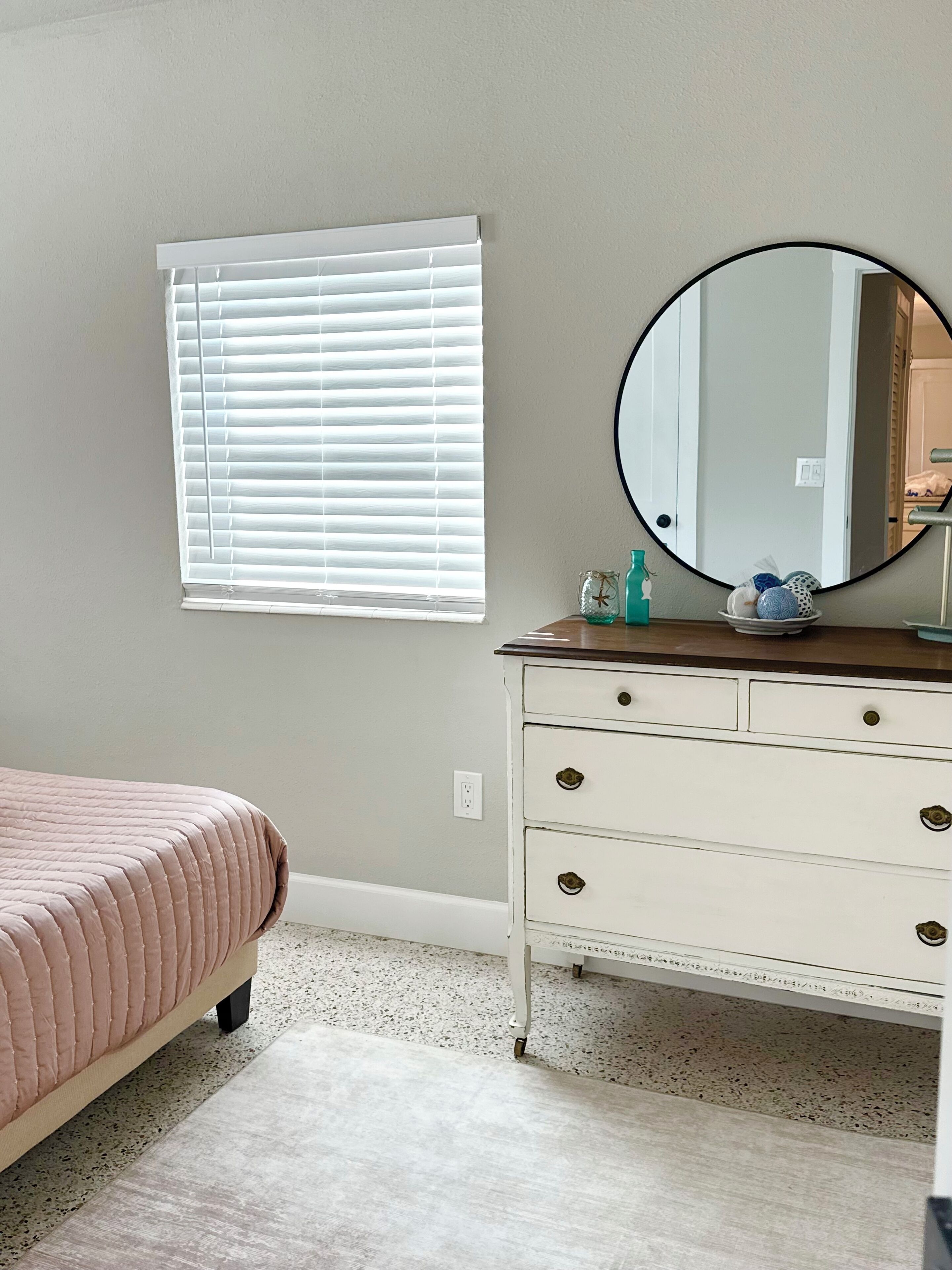 Une chambre à coucher avec un lit, une commode à tiroirs, un miroir rond et une fenêtre avec des stores.