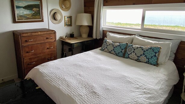 Un dormitorio con cama, cómoda, mesita de noche, lámpara, ventana con vistas y un cuadro en la pared.