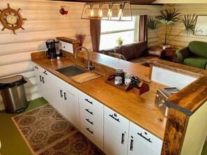 A kitchen with a wooden countertop, sink, and cabinets. There is a clock on the wall, a plant, and a window with curtains.