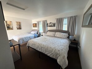 Une chambre avec deux lits simples, un bureau et une chaise. Il y a deux fenêtres avec des rideaux, une œuvre d’art accrochée au mur et un miroir sur le mur de droite.