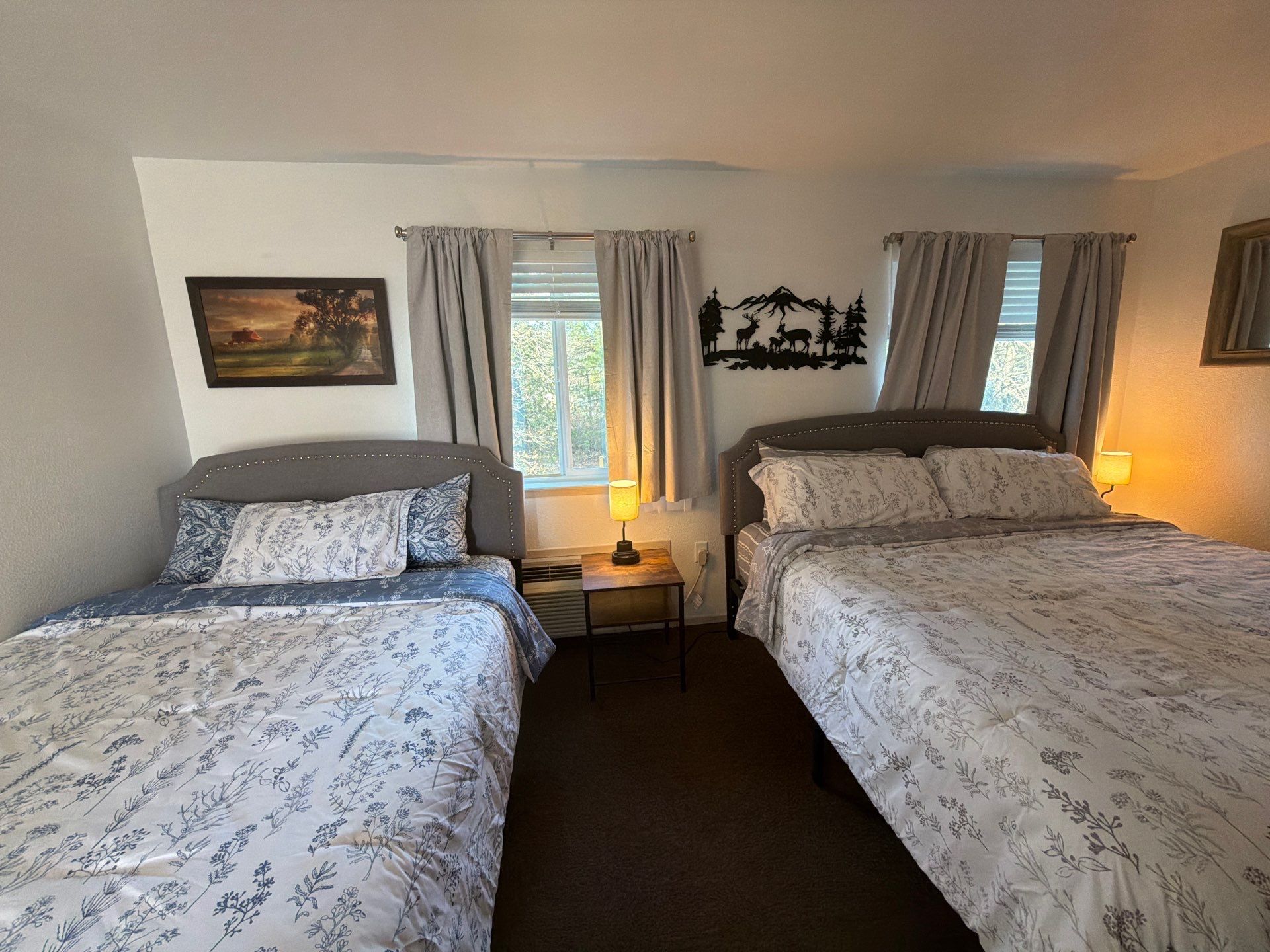 A room with two beds, each with floral-patterned bedding, a nightstand with a lamp, a painting on the wall, and a decorative wall piece.