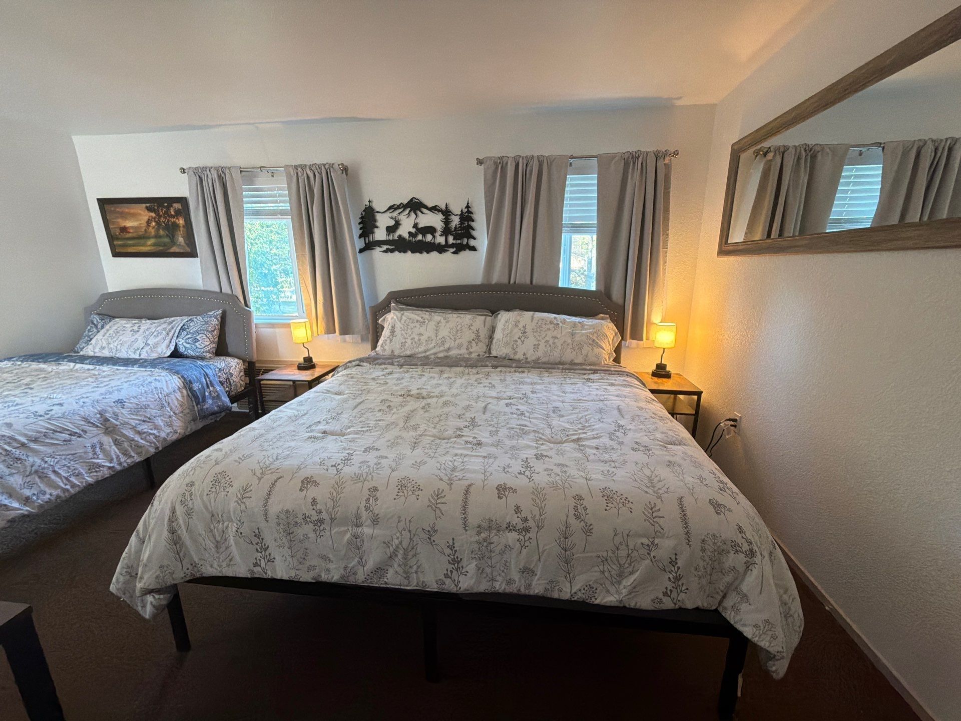 Quarto duplo com duas camas, cada uma com mesa de cabeceira e luminária. Há uma moldura com um quadro na parede acima da cama à esquerda.
