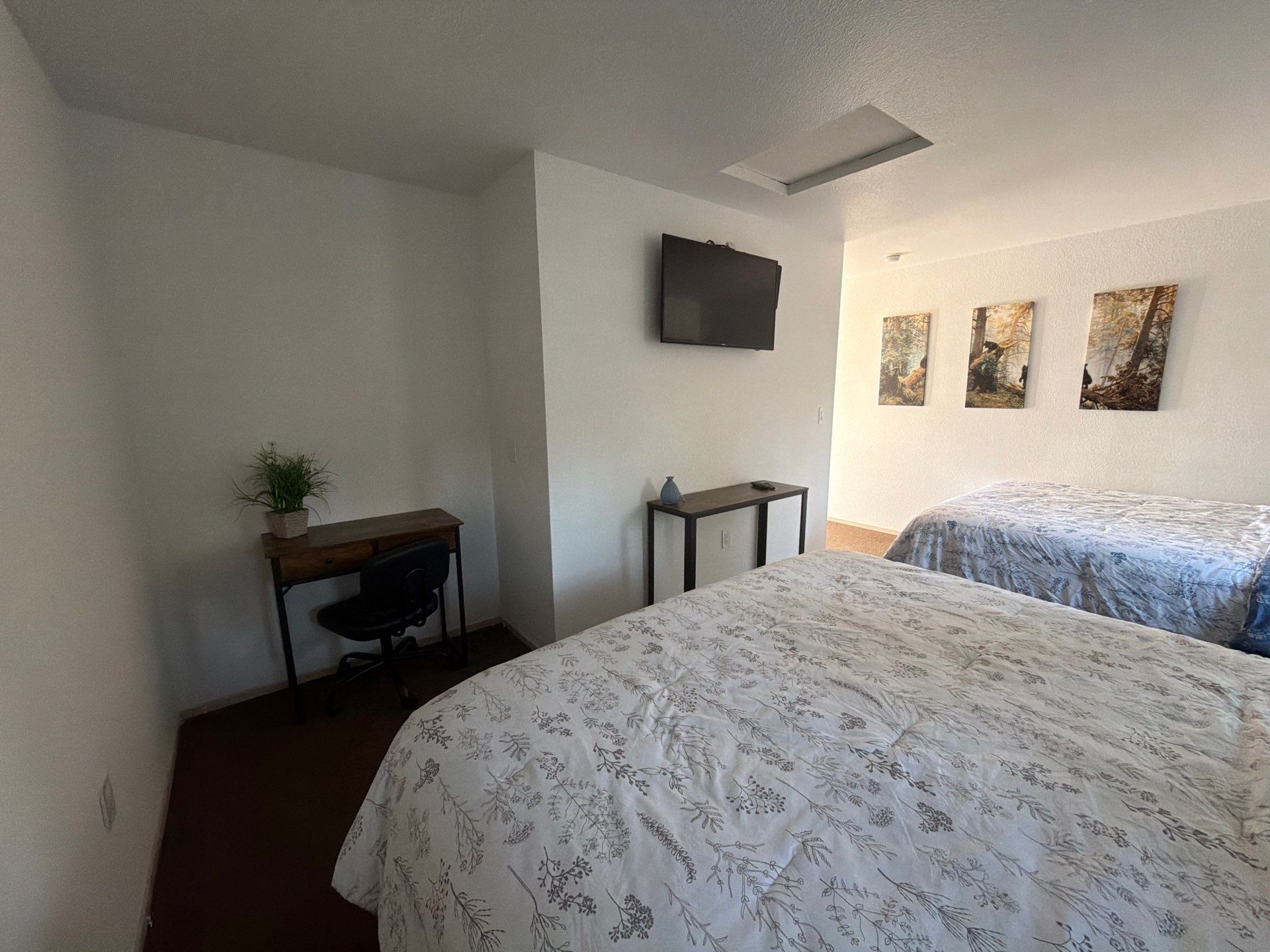 A bedroom with a bed, a desk, a chair, a small table, and a TV on the wall.