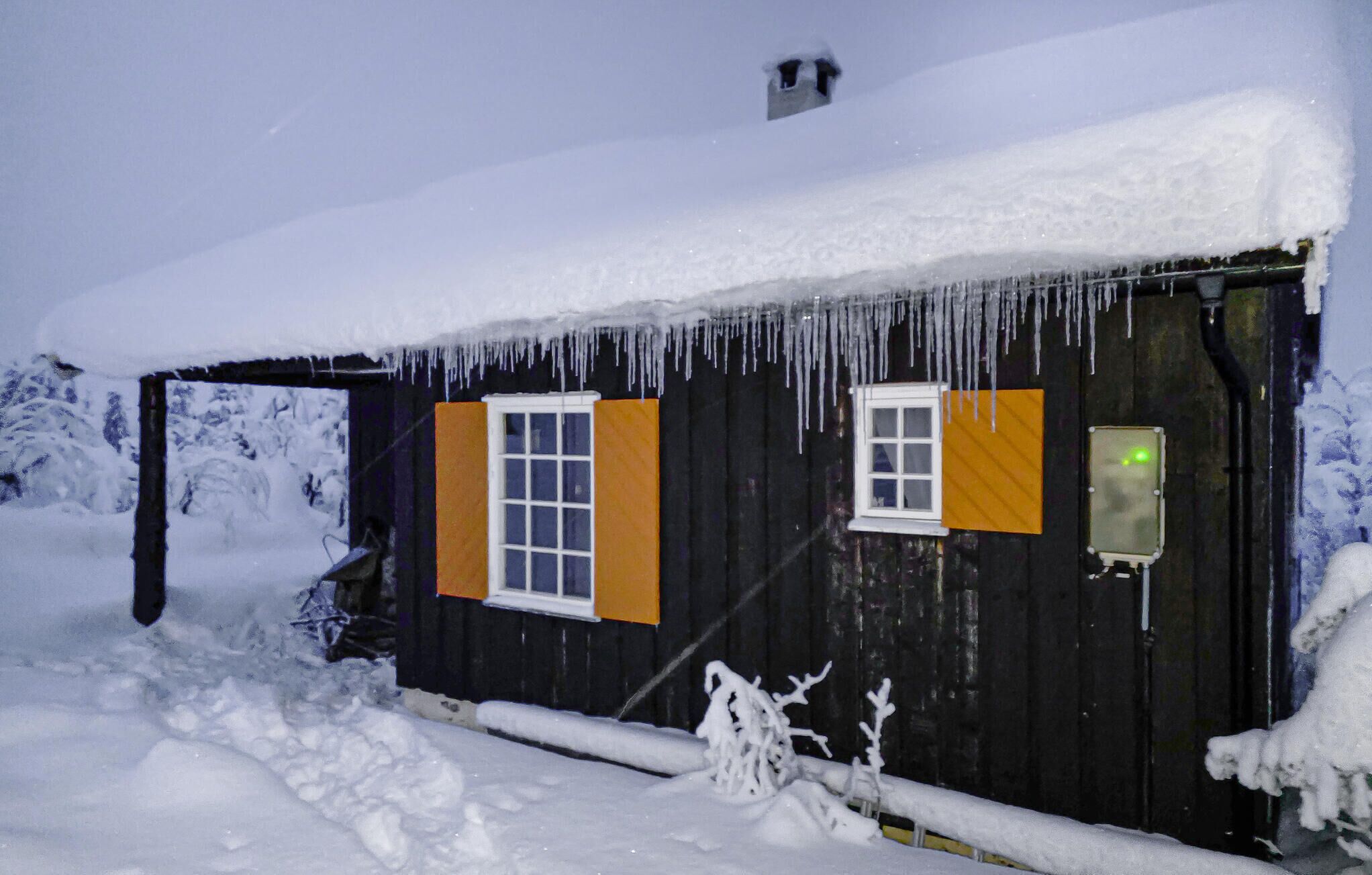 Una cabaña de madera con dos ventanas, una con contraventanas naranjas y un letrero de advertencia amarillo, rodeada de nieve.