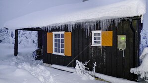 Eine Holzhütte mit zwei Fenstern, eines davon mit orangen Fensterläden und einem gelben Warnschild, umgeben von Schnee.