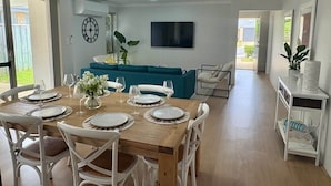 En matplats med ett träbord för fyra personer, omgiven av vita stolar. En turkosfärgad soffa står längs väggen, och en platt-TV är monterad ovanför den.