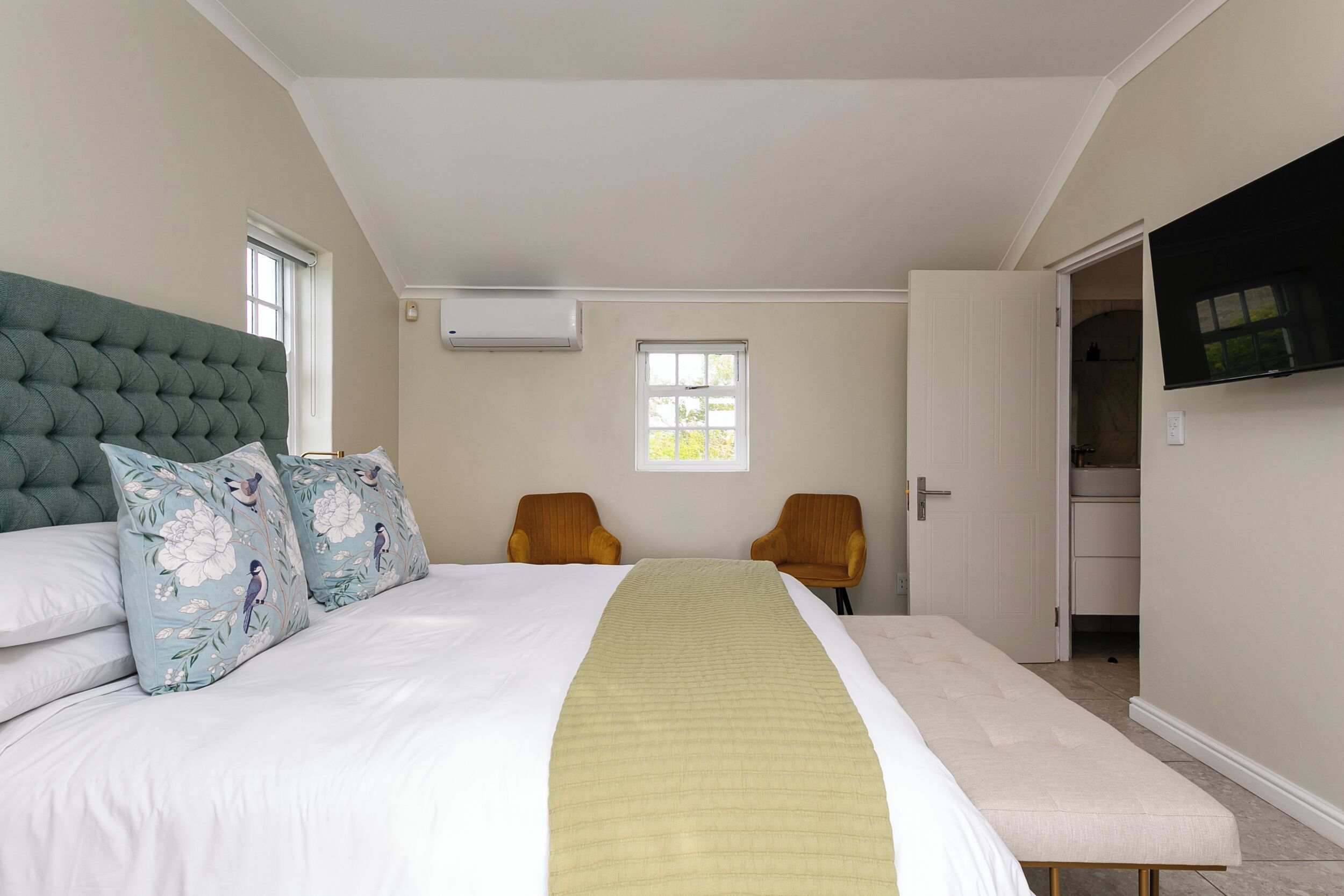 A bedroom with a large bed, two chairs, a bench, a window, and a TV mounted on the wall.