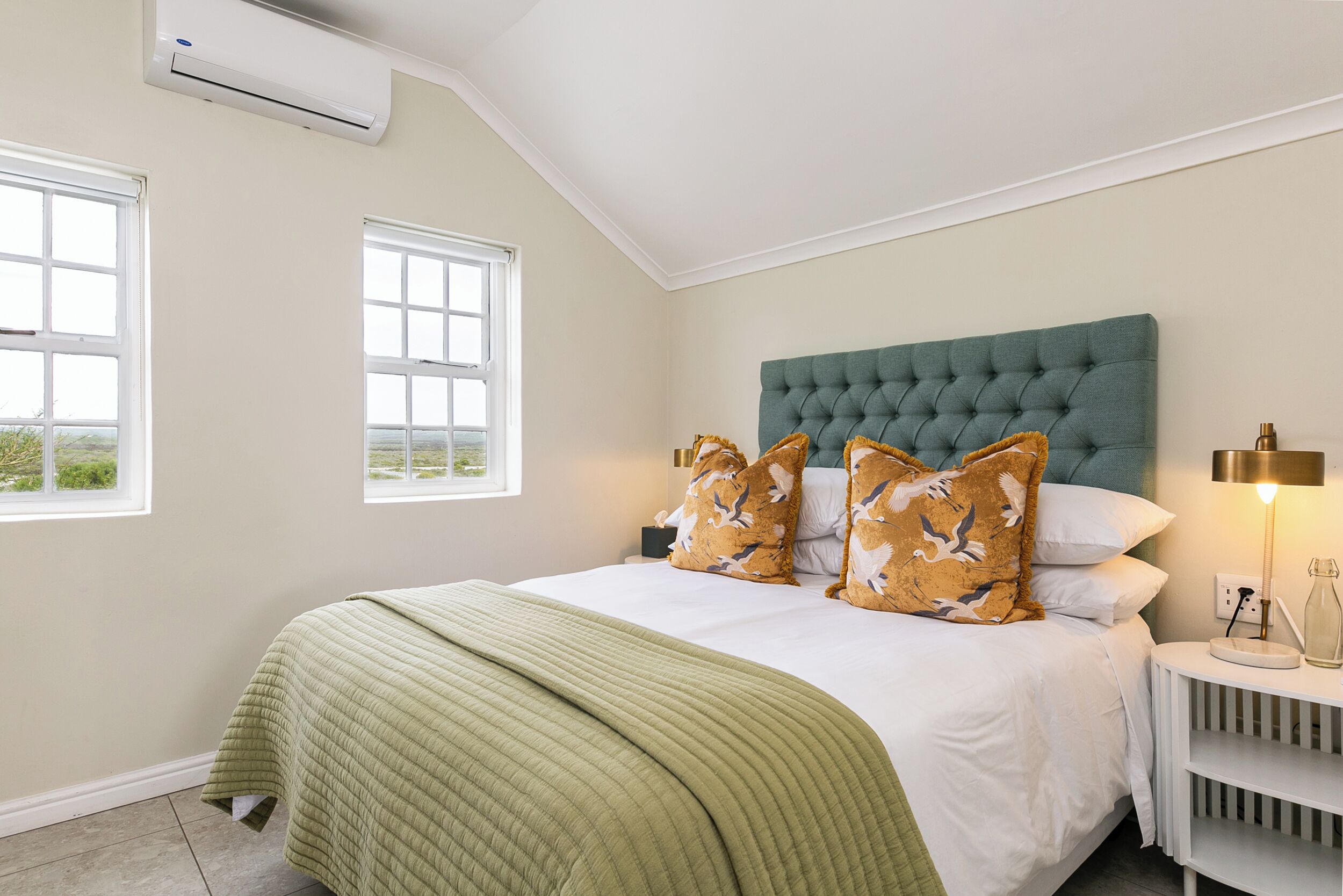 A bedroom with a large bed, two windows, a green headboard, and a bedside table with a lamp.