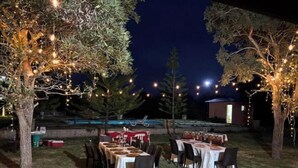 An outdoor dining area with tables set for guests, surrounded by trees and string lights.