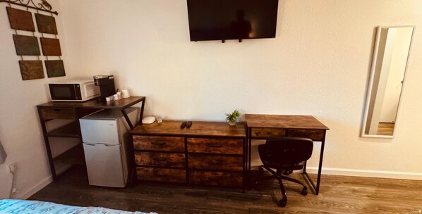 Una habitación compacta con un escritorio de madera, un refrigerador pequeño, un microondas, una silla de oficina negra y un televisor montado en la pared.