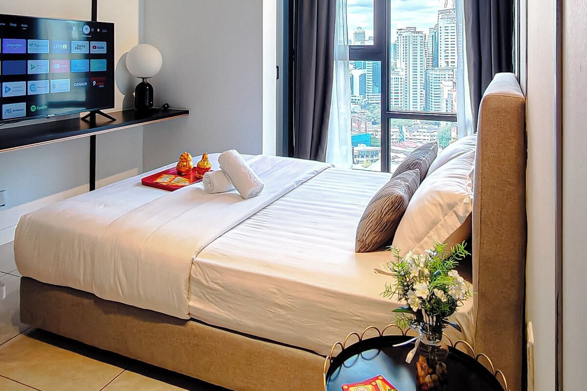A modern hotel room with a bed, a desk with a TV, a chair, and a view of a cityscape.