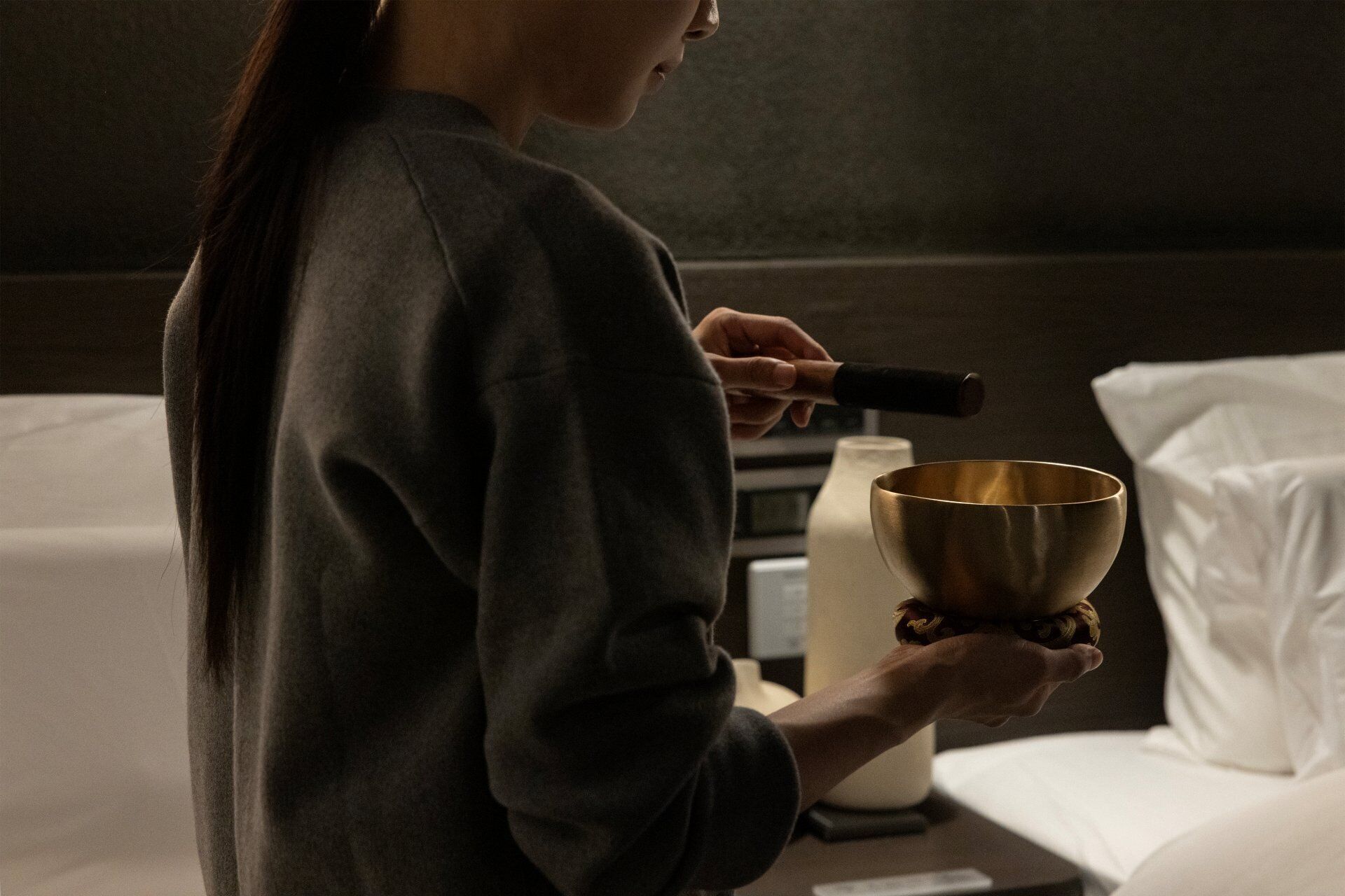 A person holding a golden bowl, standing in a room with a white bed and a white pillow.