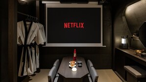 Una habitación con una pantalla de proyector mostrando “NETFLIX”, una mesa con una botella y platos, y una bata colgada en un perchero.