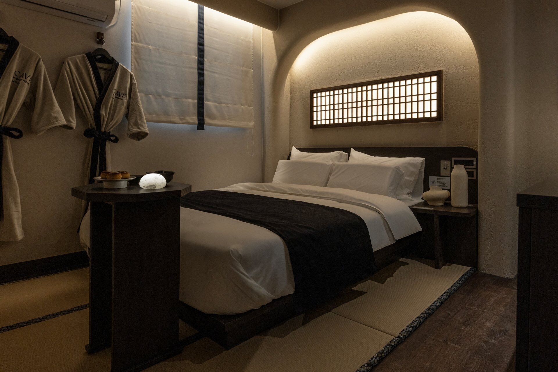 Habitación de hotel con cama, mesita de noche, lámpara de pared, perchero y ventana con vistas.