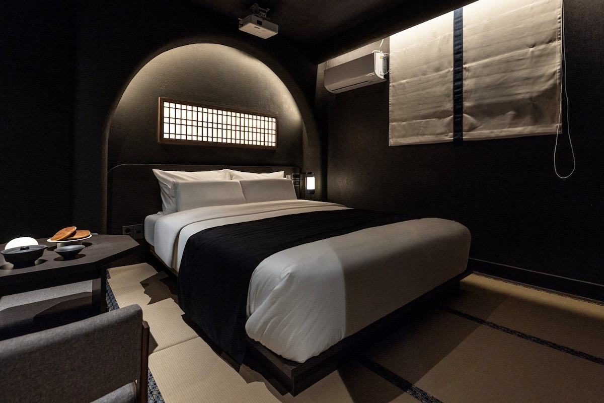 Una habitación de hotel moderna con una cama grande, una mesita con lámpara y una ventana con vistas.