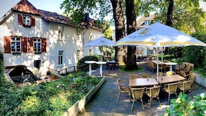 Un patio con mesas al aire libre, un edificio blanco con contraventanas de madera y un árbol grande.