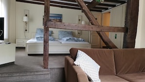 Una habitación con un sofá marrón, una cama con ropa de cama azul y blanca, una viga de techo de madera y una pequeña planta colgando del techo.