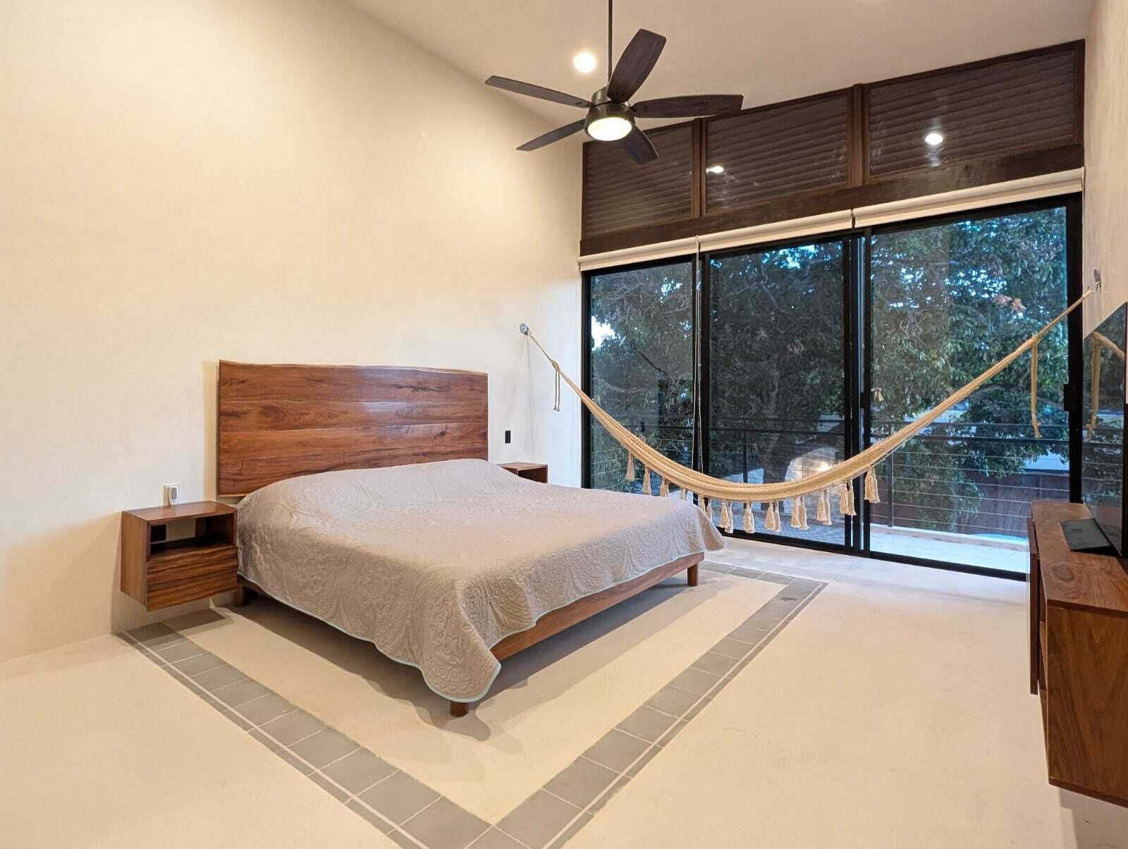 A modern bedroom with a wooden bed, a hammock, a ceiling fan, and large windows offering a view of trees.