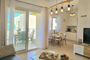 A modern living room with a sofa, a glass coffee table, a dining area with a table and chairs, and a kitchen area with cabinets and a TV. - Perla Holiday Homes 143, Lalzit Bay, Albania (Lalzit Bay)