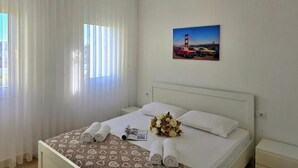 A hotel room with a bed, curtains, a wall-mounted picture, and a bedside table. - Perla Holiday Homes 143, Lalzit Bay, Albania (Lalzit Bay)