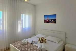 A hotel room with a bed, curtains, a wall-mounted picture, and a bedside table. - Perla Holiday Homes 143, Lalzit Bay, Albania (Lalzit Bay)