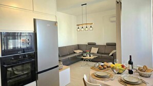 A modern kitchen with a built-in oven and refrigerator, a dining area with a round table set for four, and a grey sofa in the living area. - Perla Holiday Homes 143, Lalzit Bay, Albania (Lalzit Bay)