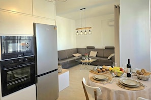 A modern kitchen with a built-in oven and refrigerator, a dining area with a round table set for four, and a grey sofa in the living area. - Perla Holiday Homes 143, Lalzit Bay, Albania (Lalzit Bay)