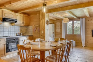 A wooden cabin interior with a kitchen, dining table, chairs, and a large window. - Hostcare - Chalet des Pistes - Nature et Détente (Crest-Voland)