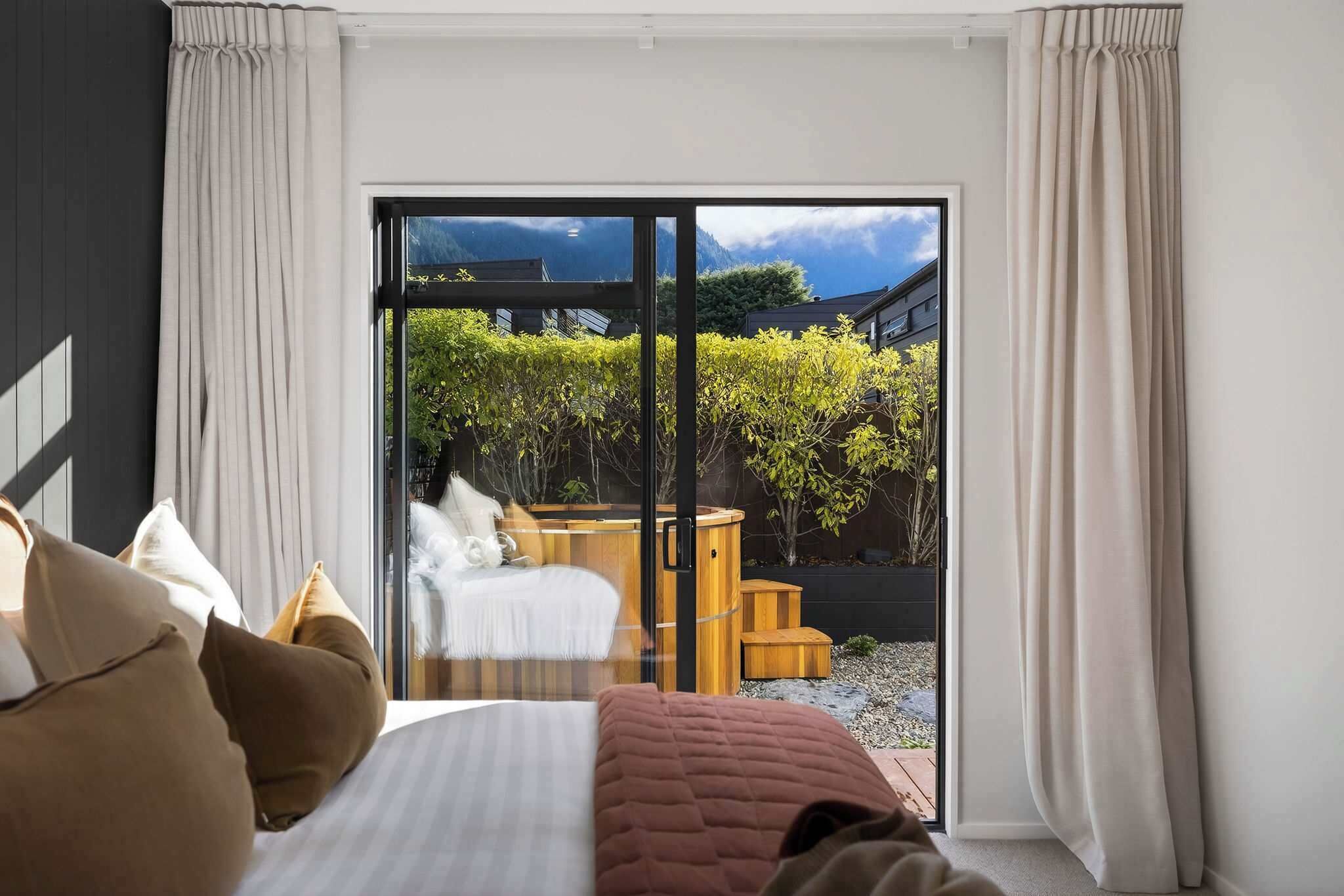 Een slaapkamer met een bed, kussens en uitzicht door een schuifdeur op een buitenruimte met een hottub en groen.