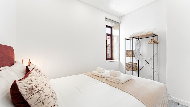 Ein schlichter Schlafzimmerraum mit einem großen Bett, einem Fenster mit Ausblick und einem Metallregal mit Körben.