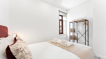 Ein schlichter Schlafzimmerraum mit einem großen Bett, einem Fenster mit Ausblick und einem Metallregal mit Körben.